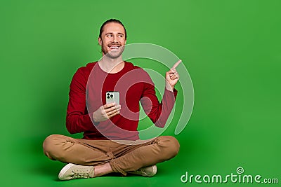 Full size photo optimistic guy wear red shirt sit with smartphone look directing at promo empty space isolated on green Stock Photo