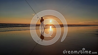 From full sun at sunset silhouetted fisherman with his very long rod on reflecting wet beach sand Stock Photo