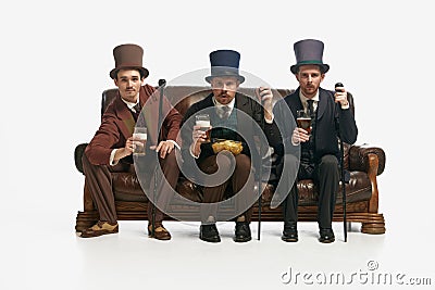 Portrait with three men wearing old-fashioned clothes sitting on sofa with snack and tense face over white background Stock Photo