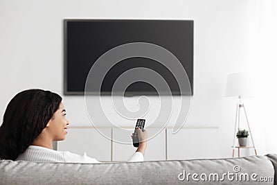 Fun in the evening. Relaxed black lady watching TV pointing remote control at flatscreen television with black screen Stock Photo