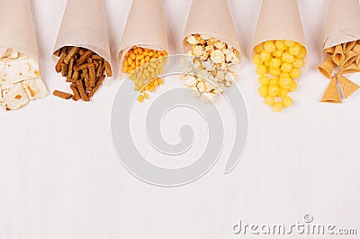 Fun fresh summer fast food background - snacks nacho, croutons, chips, tortilla, popcorn in craft paper cone on white wood board. Stock Photo