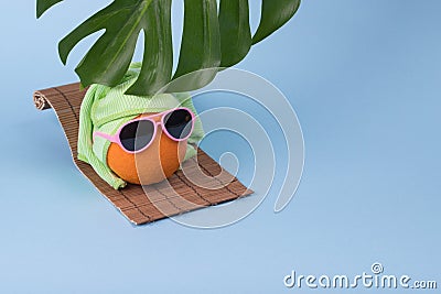 Fun idea made with an orange in sunglasses wearing a towel on his head lying on a sunbed and a monstera leaf on a blue background Stock Photo
