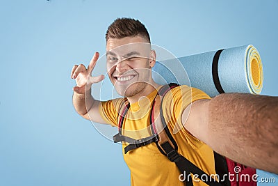 Funky millennial hipster with camping equipment taking selfie, winking and showing peace gesture over blue background Stock Photo