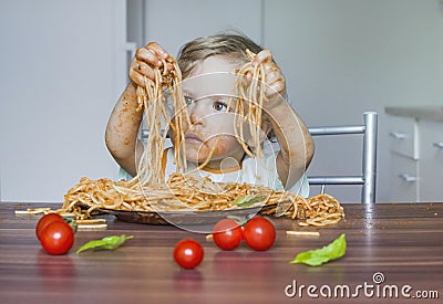 Funny baby child getting messy eating spaghetti with tomato sauce from a large plate, by itself with his hands, at home Stock Photo