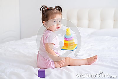 funny baby girl in pink bodysuit playing with plastic toy children pyramid on bed Stock Photo