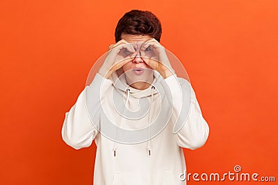 Funny curious young man in casual white sweatshirt looking far away through binoculars gesture, spying with open mouth, shocking Stock Photo