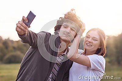 Funny curly male youngster and his girlfriend pose for making selfie portrait against blurred nature background, have positive exp Stock Photo