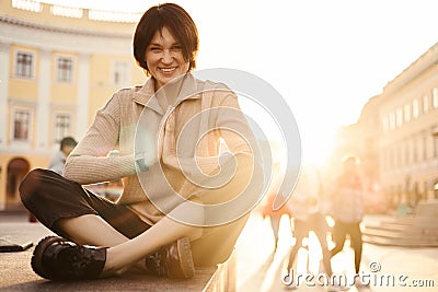 Funny fair-skinned adult sits outdoors, holding palms together and legs crossed, smiling at camera. Stock Photo