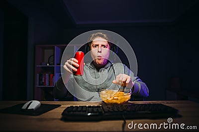 Funny gamer with a shocked face looks puzzled at the camera and eats chips from a plate and drinks cola from a red can. Guy plays Stock Photo