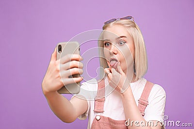 Funny girl with light hair doing a selfie on a purple background and making a funny face. A funny teen girl takes a selfie on a Stock Photo