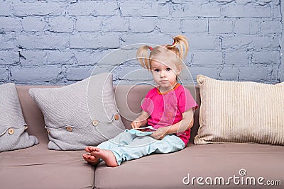 A funny girl of two years from birth with white hair and in a red shirt and blue pants plays a mobile phone on the couch in a room Stock Photo