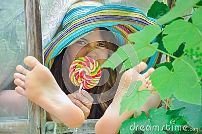 Funny girl wearing a colorful hat with lollipop and showing her feet in window with grape leaves Stock Photo