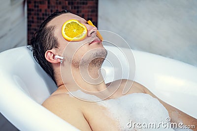 Funny guy washing his bath. Listens to music with wireless headphones. Relaxed from spa procedures on face with orange round slice Stock Photo