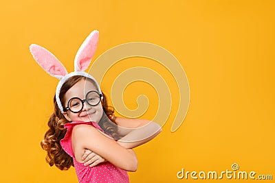 Funny happy cute little girl wearing Easter bunny ears and glasses on a yellow background. The child hugs himself with his arms Stock Photo