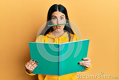 Funny hispanic woman reading a book doing crazy gesture with pencil over mouth like mustache, playful and positive on free time, Stock Photo