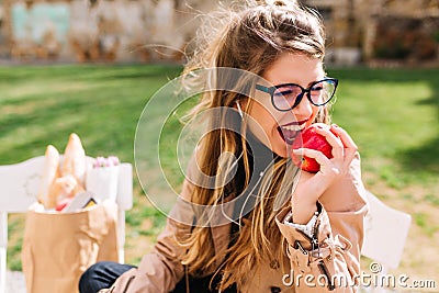 Funny hungry girl with appetite bites off red apple sitting in the park after a shopping trip. Attractive young woman Stock Photo