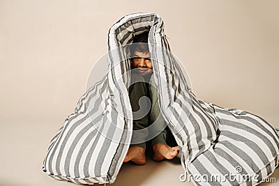 Funny indian boy sitting under a striped mattress over beige background. Stock Photo