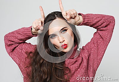 Funny joking young woman, feels happy, makes horns on her head with fingers, wears pink sweater Stock Photo