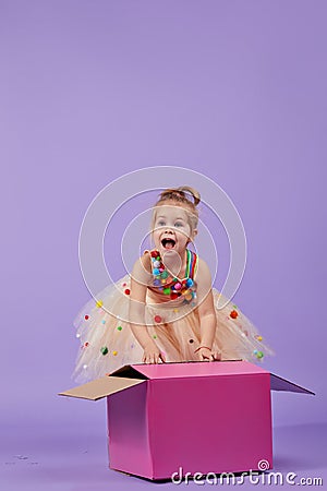 A funny little girl child in a magnificent holiday dress look inside a large gift colored box, Stock Photo