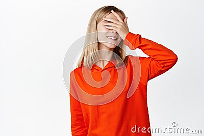 Funny little girl waits for surprise with closed eyes, cover with palm and smiling, standing over white background Stock Photo