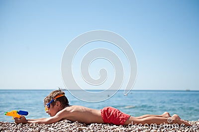 Funny little lying caucasian boy with water toy gun and goggles aiming on summer sea beach during travel vacation Stock Photo