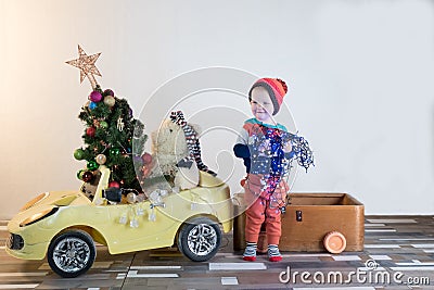 Funny little smiling kids driving toy car with Christmas tree. Happy child in colour fashion clothes bringing hewed xmas tree from Stock Photo