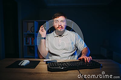 Funny man with a beard smokes ikos at home at work on the computer at night and looks into the camera with a surprised face Stock Photo