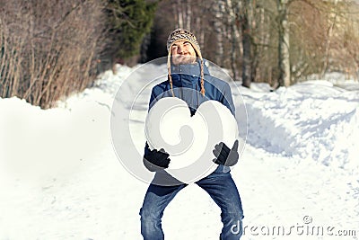 Funny man with great difficulty holding huge heart made of snow, standing on snowy road in winter park. Fall in love. Declarations Stock Photo