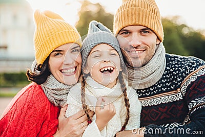 Funny overjoyed small kid laughs as has wonderful time with her parents. Affectionate parents stand near their little daughter who Stock Photo