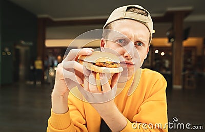Funny photo of a cheerful young man in a cap and a yellow sweater sits in a fast-food restaurant, looks at the camera and makes a Stock Photo
