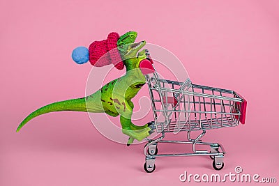 Plastic toy dinosaur wearing tiny knitted hat and driving empty shopping trolley on a pink background ,shopping concept Editorial Stock Photo