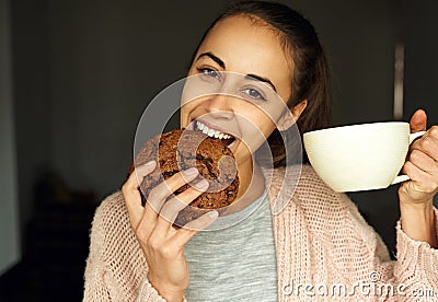 Funny portrait of happy young woman with large cup tea, biting huge piece of chocolate cookie. Stock Photo
