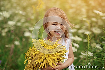 Funny red-haired girl laughs merrily looking into the camera with a bouquet of yellow flowers in her hands in a field at sunset. Stock Photo