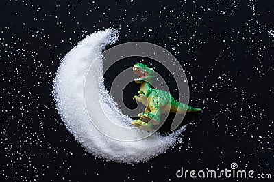 Funny toy dinosaur stands on a white powder crescent moon. Black background. Fabulous concept of the theory of the origin and Stock Photo