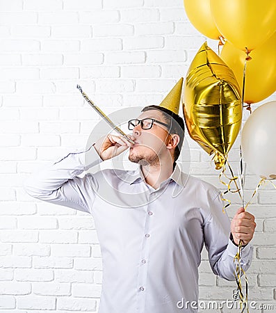 Funny young man wearing birthday hat and holding balloons blowing noisemaker celebrating birthday Stock Photo