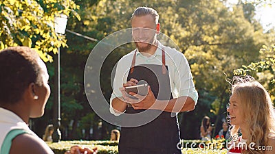 A further back view of a beautiful park with lots of tables, a sexy waiter with a beard and blonde hair is taking orders Stock Photo