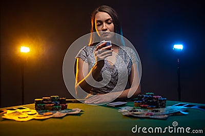 Gambler woman siting at the table in casino with poker face. Lucky player on smoky background with lamps. Concept of poker game Stock Photo