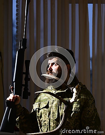 Gamekeeper concept. Man with beard wears camouflage clothing, curtains background. Macho on strict face at gamekeepers Stock Photo