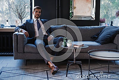 Young and charming businessman is thinking about business while sitting on the sofa in his modern office. Stock Photo