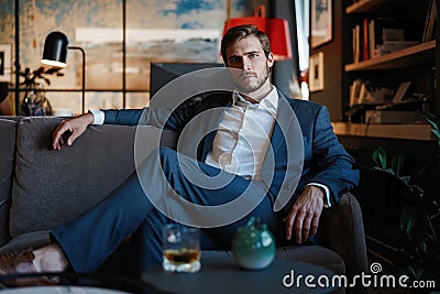 Young and charming businessman is thinking about business while sitting on the sofa in his modern office. Stock Photo