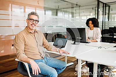 A genial senior business leader sits at an office table with a laptop Stock Photo