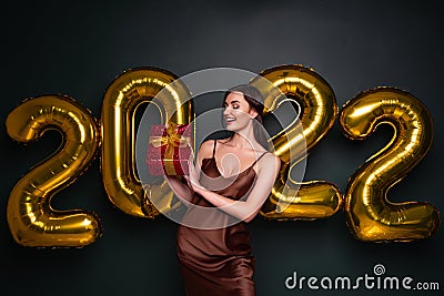 Georgeous brunette lady in beautiful dress holding gift box on black studio background with golden 2022 balloons Stock Photo