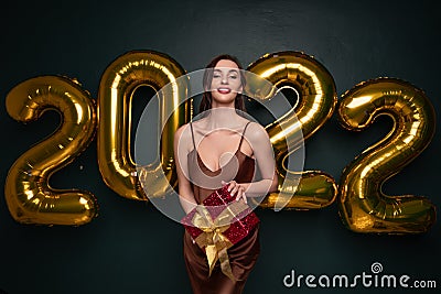 Georgeous brunette lady in beautiful dress holding gift box on black studio background with golden 2022 balloons Stock Photo
