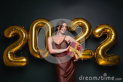 Georgeous brunette lady in beautiful dress holding gift box on black studio background with golden 2022 balloons Stock Photo