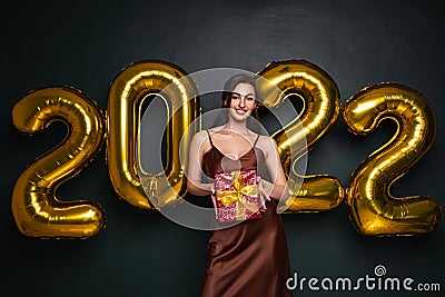 Georgeous brunette lady in beautiful dress holding gift box on black studio background with golden 2022 balloons Stock Photo