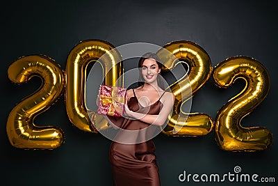 Georgeous brunette lady in beautiful dress holding gift box on black studio background with golden 2022 balloons Stock Photo