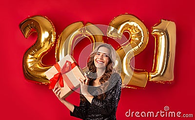 Georgeous millennial lady in beautiful dress holding gift box on red studio background with golden 2021 balloons Stock Photo