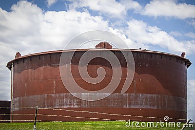 Giant rusty oil tank in tank farm in Cushing Oklahoma behind barbed wire fence with pretty blue sky background Stock Photo