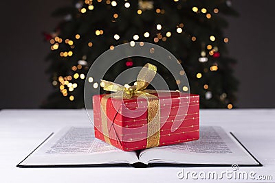 Gift. Box in red packaging. Golden bow. A gift on a book of the Bible. On the background of the Christmas tree Stock Photo