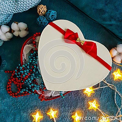 A gift box in the shape of a heart with a red bow against the background of cozy turquoise blankets framed in decorative cotton, Stock Photo